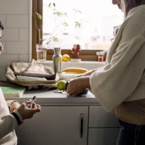 Two people in a kitchen, one holding a peeled fruit and the other a small device. A window with plants and outdoor view in the background.